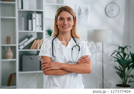 Standing with arms crossed. Young female doctor in white coat is indoors Standing with arms crossed. Young female doctor in white coat is indoors 127377404