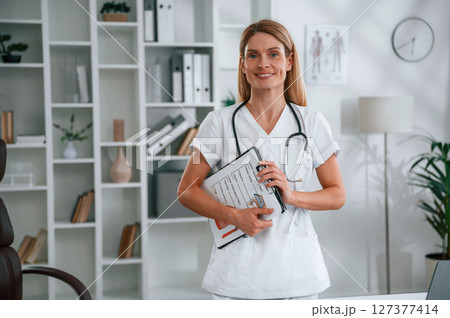 With documents in hands. Young female doctor in white coat is indoors 127377414