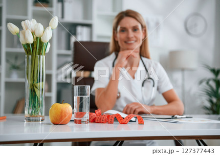 Measuring tape, flowers, an apple and glass of water are on the table. Young female doctor in white coat is indoors 127377443