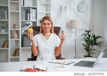 Holding an apple and glass of water. Young female doctor in white coat is indoors Holding an apple and glass of water. Young female doctor in white coat is indoors 127377451