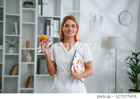 Conception of diet, healthy food. Holding an apple. Young female doctor in white coat is indoors 127377458