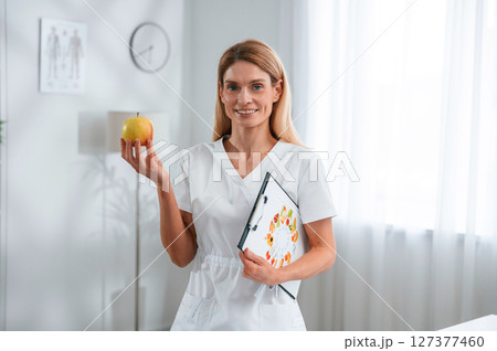 Conception of diet, healthy food. Holding an apple. Young female doctor in white coat is indoors Conception of diet, healthy food. Holding an apple. Young female doctor in white coat is indoors 127377460