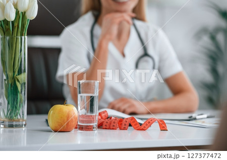 Measuring tape, an apple and glass of water are on the table. Young female doctor in white coat is indoors Measuring tape, an apple and glass of water are on the table. Young female doctor in white coat is indoors 127377472