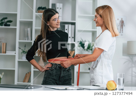 Standing and measuring the waist of customer. Young woman at a nutritionist's appointment. Medical care Standing and measuring the waist of customer. Young woman at a nutritionist's appointment. Medical care 127377494