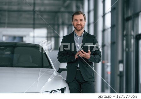 With tablet in hands. Man in black suit is indoors in the car dealership near automobile 127377658