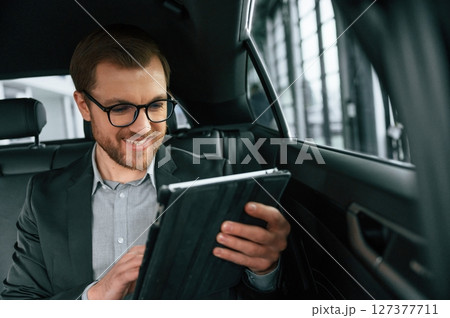 Smiling, using tablet. Man in formal business clothes is sitting in the modern automobile 127377711