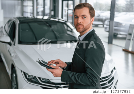 Standing, side view, holding tablet. Man in black suit is indoors in the car dealership near automobile Standing, side view, holding tablet. Man in black suit is indoors in the car dealership near automobile 127377952