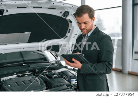 Standing, side view, holding tablet. Man in black suit is indoors in the car dealership near automobile Standing, side view, holding tablet. Man in black suit is indoors in the car dealership near automobile 127377954