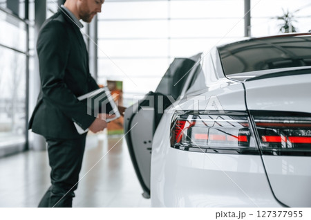Man in black suit is indoors in the car dealership near automobile Man in black suit is indoors in the car dealership near automobile 127377955