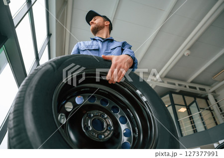 With big new tire for a change. Man in blue uniform is working in the car service With big new tire for a change. Man in blue uniform is working in the car service 127377991