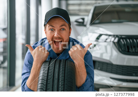 Leaning on the tire and smiling. Man in blue uniform is working in the car service 127377998