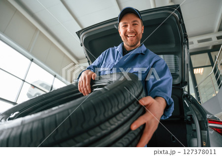 Putting the tire out of automobile. Man in blue uniform is working in the car service Putting the tire out of automobile. Man in blue uniform is working in the car service 127378011
