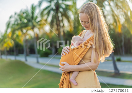 Mother holding her little baby in a yellow sling in the park. Warm and loving family moment. Babywearing, parenting, and mother-child bonding concept Mother holding her little baby in a yellow sling in the park. Warm and loving family moment. Babywearing, parenting, and mother-child bonding concept 127378031