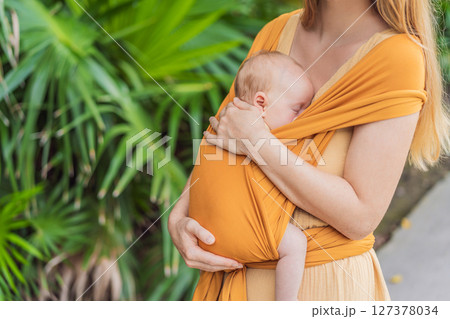Mother holding her little baby in a yellow sling in the park. Warm and loving family moment. Babywearing, parenting, and mother-child bonding concept Mother holding her little baby in a yellow sling in the park. Warm and loving family moment. Babywearing, parenting, and mother-child bonding concept 127378034