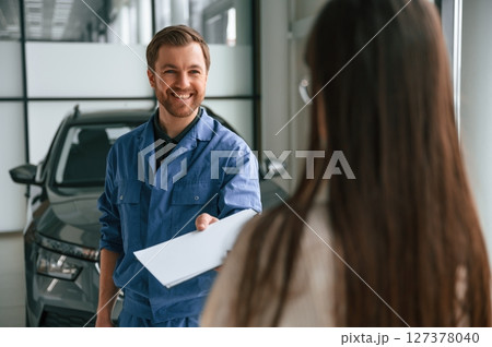 Man giving the document. Young woman is visiting auto mechanic in garage. Repair service Man giving the document. Young woman is visiting auto mechanic in garage. Repair service 127378040