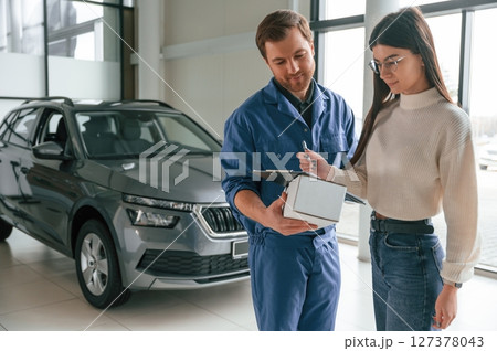 Man is holding box and notepad. Young woman is visiting auto mechanic in garage. Repair service 127378043