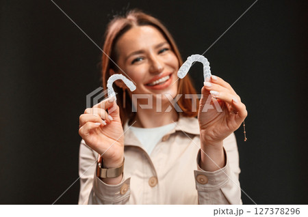 Smiling, and holding two aligners. Young woman is standing against black background Smiling, and holding two aligners. Young woman is standing against black background 127378296