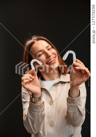 Smiling, and holding two aligners. Young woman is standing against black background Smiling, and holding two aligners. Young woman is standing against black background 127378298