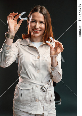 Positive young woman is standing against black background and holding aligner 127378315