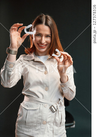 Positive young woman is standing against black background and holding aligner Positive young woman is standing against black background and holding aligner 127378316
