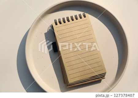 Minimalist top-down still life. Brown empty spiral notebook mockup with lined pages placed on ceramic plate, soft natural light. Creative, conceptual arrangement. Planning concept. Flat lay, top view. 127378407
