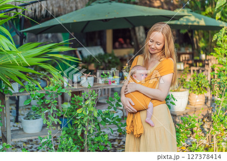 Mother holding her little baby in a yellow sling in the park. Warm and loving family moment. Babywearing, parenting, and mother-child bonding concept 127378414