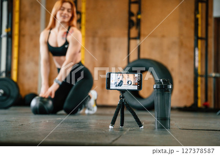 Smartphone is on the tripod, live stream, capturing video. Beautiful strong woman is in the gym Smartphone is on the tripod, live stream, capturing video. Beautiful strong woman is in the gym 127378587