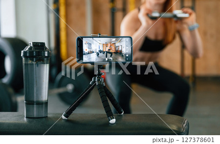 Doing squats, shooting video by smartphone. Beautiful strong woman is in the gym 127378601