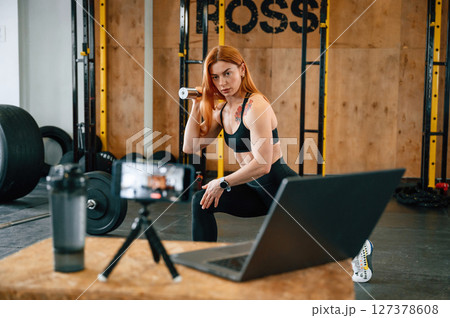 Laptop, bottle and smartphone on tripod is on the table. Beautiful strong woman is in the gym Laptop, bottle and smartphone on tripod is on the table. Beautiful strong woman is in the gym 127378608