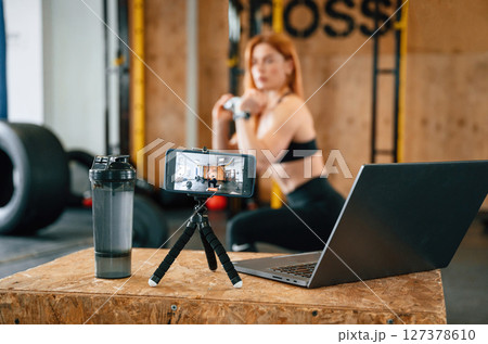 Laptop, bottle and smartphone on tripod is on the table. Beautiful strong woman is in the gym 127378610