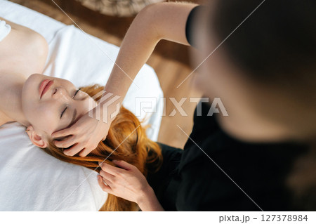 Top view of professional masseuse gently massaging temples of relaxed redhead woman with closed eyes, providing soothing head massage on comfortable massage table in spa or wellness center, close-up. 127378984