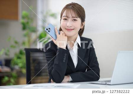 スマホを持つディスクワーク中の若いビジネスウーマン スマホを持つディスクワーク中の若いビジネスウーマン 127382222