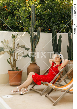 Relaxed woman in red dress sunbathing in lounge chair by cactus garden wall on a summer day 127385041