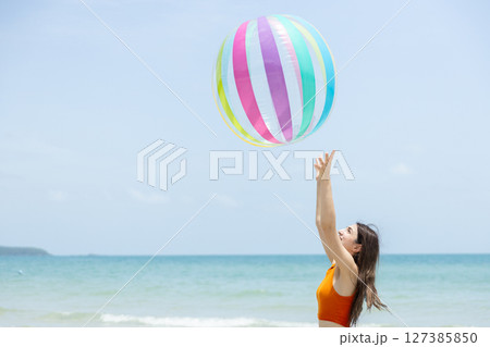 beautiful brunette women wearing white shorts and orange top tossing large, colorful beach ball into air at beach on bright day. Relaxation happy holiday. beautiful brunette women wearing white shorts and orange top tossing large, colorful beach ball into air at beach on bright day. Relaxation happy holiday. 127385850