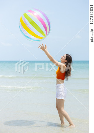 beautiful brunette women wearing white shorts and orange top tossing large, colorful beach ball into air at beach on bright day. Relaxation happy holiday. beautiful brunette women wearing white shorts and orange top tossing large, colorful beach ball into air at beach on bright day. Relaxation happy holiday. 127385851