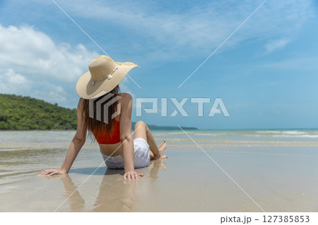 Women with straw hat, orange top and white shorts sitting on beach in tropical location with blue sky and turquoise sea. Back view. Women with straw hat, orange top and white shorts sitting on beach in tropical location with blue sky and turquoise sea. Back view. 127385853