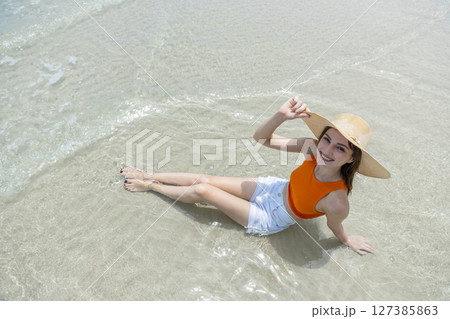 Young smiling women with straw hat, orange top and white shorts sitting on beach in tropical location with blue sky and turquoise sea. 127385863