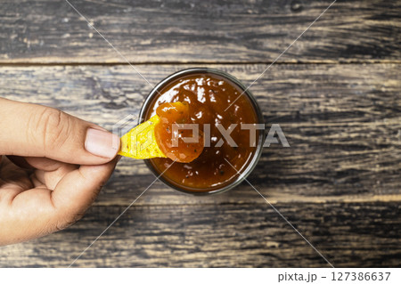 Human hand dipping a nachos tortilla chip in salsa barbecue sauce in a bowl on the table. Food and snacks concept Human hand dipping a nachos tortilla chip in salsa barbecue sauce in a bowl on the table. Food and snacks concept 127386637