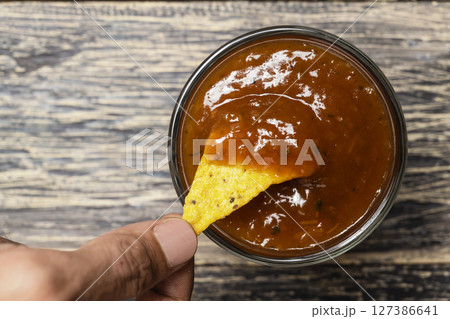 Human hand dipping a nachos tortilla chip in salsa barbecue sauce in a bowl on the table. Food and snacks concept Human hand dipping a nachos tortilla chip in salsa barbecue sauce in a bowl on the table. Food and snacks concept 127386641