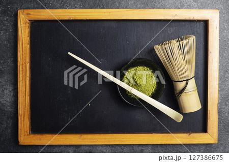 Matcha green tea powder in the black bowl and a traditional bamboo brush whisk with a spoon on a black table. Accessories for making matcha green tea. Copy space. Top view 127386675
