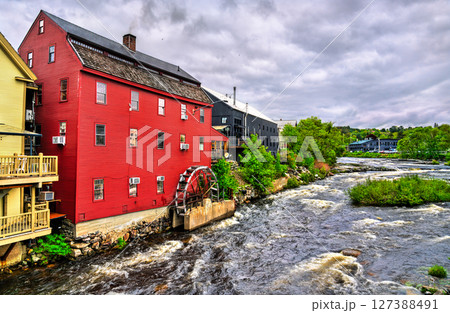 In Littleton, New Hampshire, United States, a red clapboard grist mill with a working waterwheel stands above the rushing Ammonoosuc River beneath a bright, cloud-dappled summer sky. 127388491