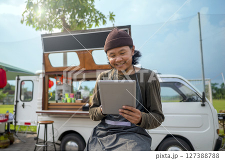 Indonesian southeast asian male barista sitting while using a tablet in front of his coffee shop. A small business of a coffee shop or cafe on a truck Indonesian southeast asian male barista sitting while using a tablet in front of his coffee shop. A small business of a coffee shop or cafe on a truck 127388788