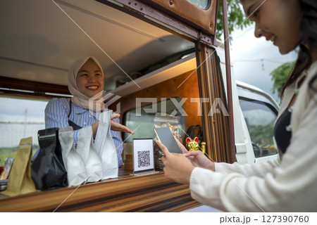 Chinese muslim female barista in hijab showing qr code to indonesian southeast asian woman to scan with cellphone for payment. Mockup or copy space. Small business of coffee shop or cafe on truck 127390760