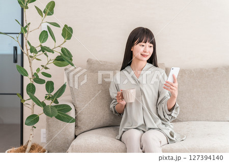 自宅のソファに座りコーヒーを飲みながらスマホを見る若いアジア人女性 127394140