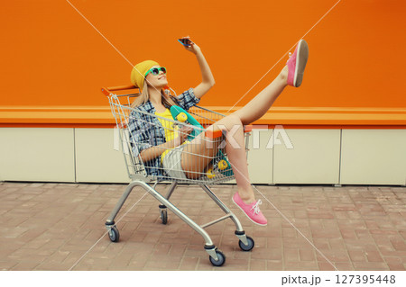 Stylish young woman taking selfie with phone, cool teenage girl in shopping trolley cart in the city 127395448