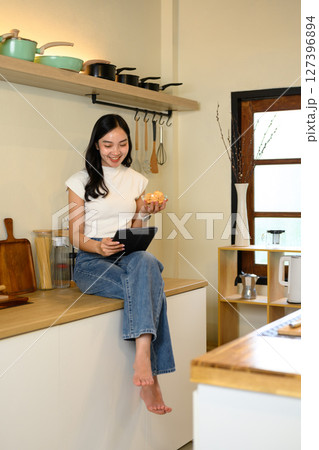 Relaxed young woman sitting on a kitchen counter, eating fresh apple and using a digital tablet Relaxed young woman sitting on a kitchen counter, eating fresh apple and using a digital tablet 127396894