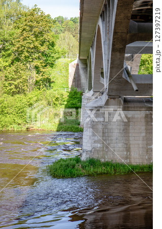 Concrete Bridge Structure Over Flowing River with Green Trees and Reflections in Daylight Concrete Bridge Structure Over Flowing River with Green Trees and Reflections in Daylight 127397219