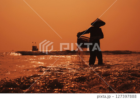 Silhouette of a Vietnamese woman in straw hat on shore of sea with ships in fishing village at sunset in Vietnam 127397888