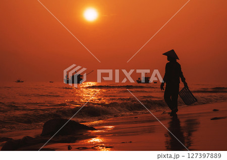 Silhouette of a Vietnamese woman in a straw hat on shore of sea with ships in a fishing village in the morning at sunrise in Vietnam Silhouette of a Vietnamese woman in a straw hat on shore of sea with ships in a fishing village in the morning at sunrise in Vietnam 127397889