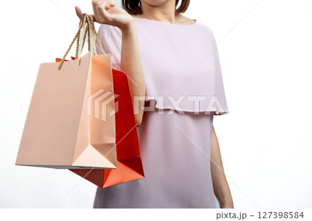 Woman in pastel pink dress holding orange and peach shopping bags with golden rope handles on her arm, isolated on white background; concept of retail or lifestyle spending Woman in pastel pink dress holding orange and peach shopping bags with golden rope handles on her arm, isolated on white background; concept of retail or lifestyle spending 127398584